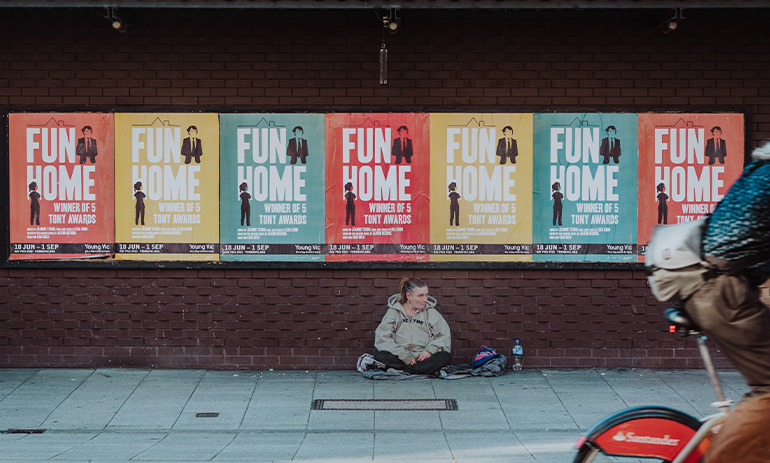 A woman sits on a street path beneath colourful posters as a bike rides past. It is apparent that she is experiencing homelessness.