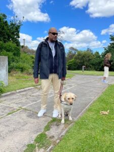 Antony, wearing a blue jacket and beige pants, stands with guide dog graduate Brett in a garden.