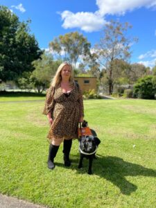 Nikki, in a leopard print dress, stands with her black guide dog Perdie in a garden.