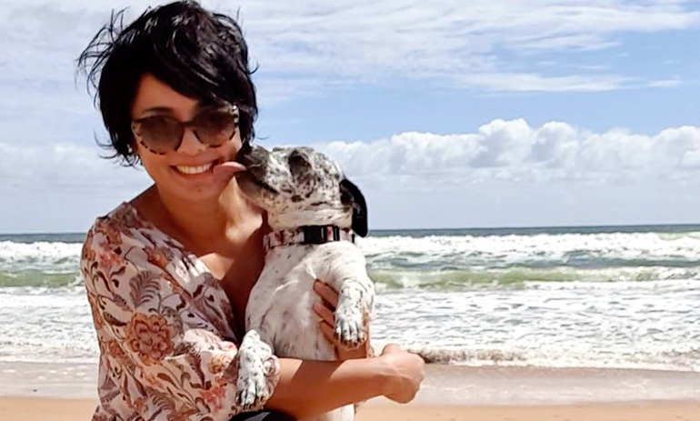 A young dark haired lady is sitting on the beach, holding a spotty black and white dog. The dog is licking her face and she is smiling. She is wearing sunglasses.