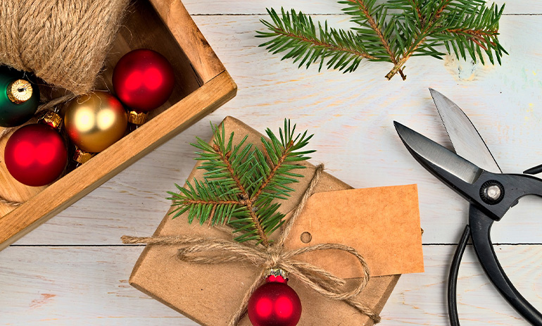 A present wrapped in brown paper and decorated with a bauble and a sprig of pine. Next to it are scissors, a box of baubles, and more sprigs of pine.