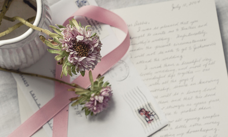 A letter in cursive writing sits open beneath a vase of flowers with a pink ribbon.