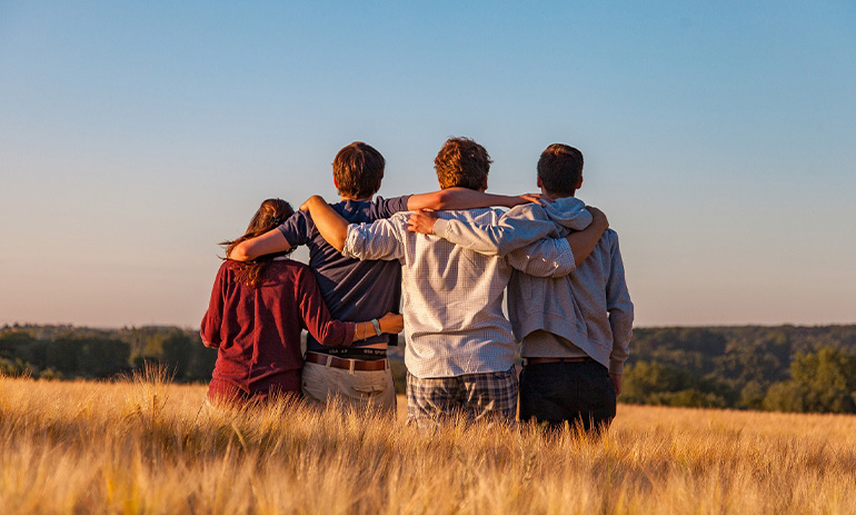 A group of young people stand in a field with their arms around each other and their backs to the camera.