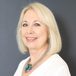 We see an older lady, her face framed by pale hair and her eyes lined. She is in a white shirt with a necklace with a blue stone in the centre. She is smiling over her shoulder.