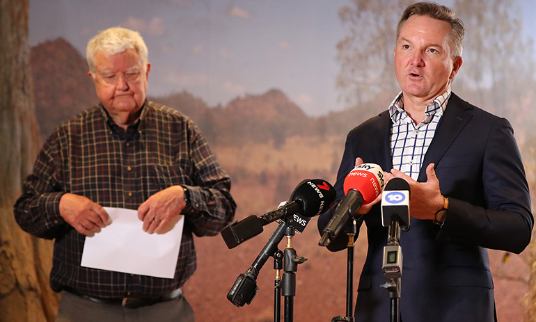 Two men stand in front of microphones before a country backdrop. One is older, with white hair and a check shirt, holding a piece of paper. The other is younger and in a suit with no tie.
