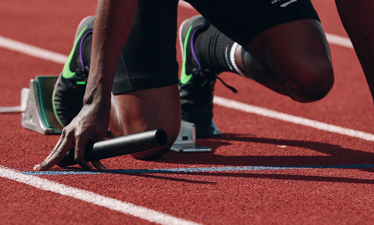 A black man kneels on a running track, holding a baton, ready to run a race.