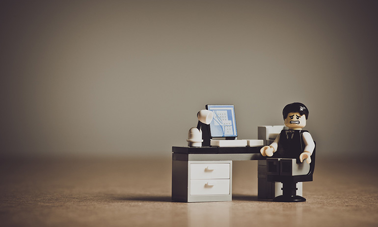 A lego figure at a mini desk with a computer screen on it. He is turned towards the camera and looks upset.