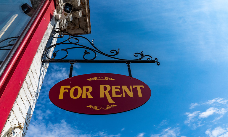 Looking up at a sign hanging off a building that says For Rent in yellow text on an ornate red oval shaped sign.