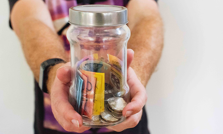 A person holds out a glass jar filled with Australian money.