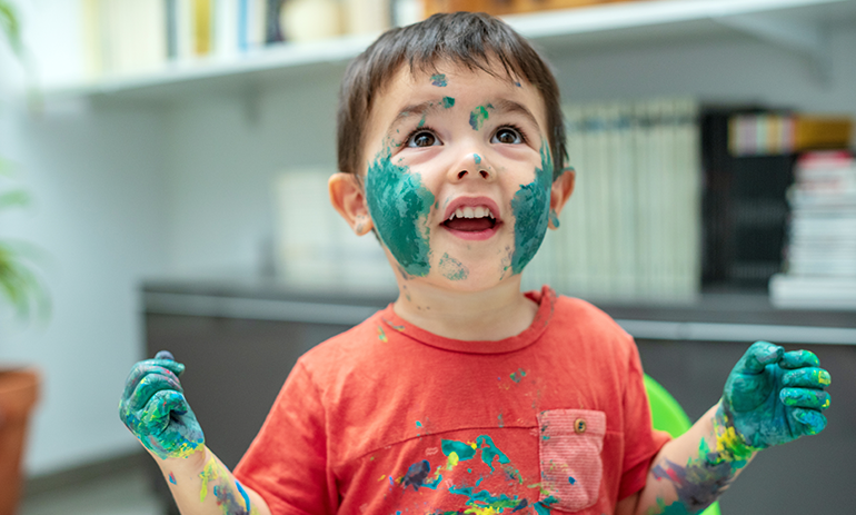 Boy toddler in orange shirt covered in paint smiling