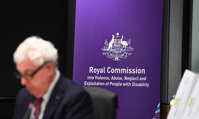 We see a white haired man in a suit in the front of the image, blurred. He is wearing black glasses. In the background behind him, in sharper focus, is a purple sign with the Australian government logo on it and the words Royal Commission into Violence, Abuse, Neglect and Exploitation of People with Disability and the commission website https://disability.royalcommission.gov.au/