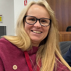 Jody Barney, a lady with long blonde hair and black rimmed glasses, smiles at the camera. She is wearing a maroon top.