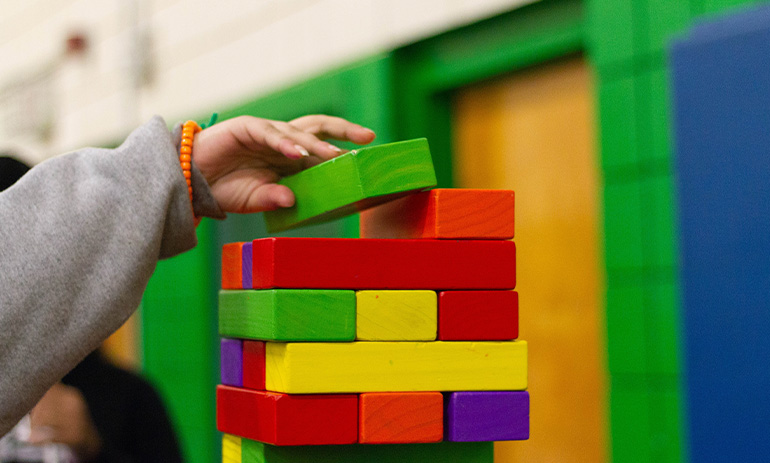 We see a child's hand reaching up to place a green block on a tower of colourful blocks. The child is wearing a grey sleeved top.