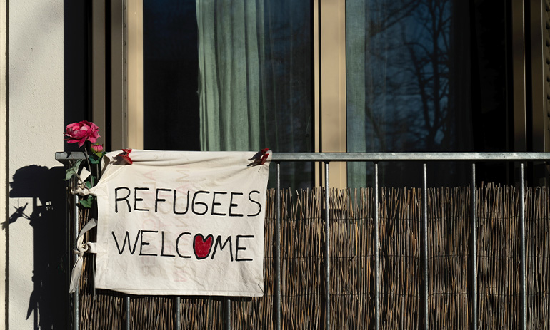 A building with a metal railing fence outside. A pink rose is tied to the fence, with a white sign that says 'refugees welcome', with a red heart.