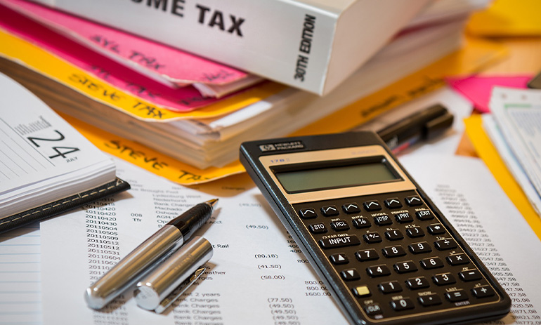 A table laid out with things to do with tax, including papers covered in columns of numbers, a black calculator, a fountain pen, a book about income tax and files that say 'tax'.