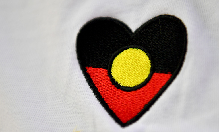The Aboriginal Flag in the shape of a heart, with black at the top, a yellow circle in the centre, and red on the bottom, against a white background.