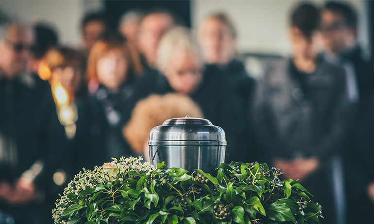 Urn of ashes sitting on bed of flowers with people dressed in black in background.