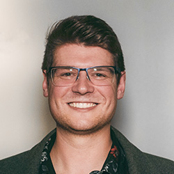 Joshua Morley, a white man, smiles at the camera in this headshot. He is wearing dark clothes and glasses, and has brown hair.