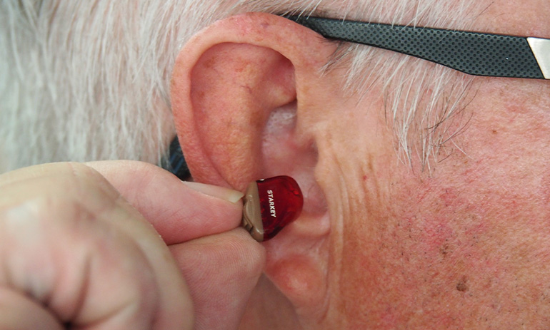 A grey haired, elderly man holds a tiny red hearing aid up by his ear. We can also see the black arm of his glasses over his ear.
