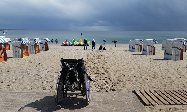 A beach scene, with huts along the sand. In the foregound is a wheelchair, facing out towards the water. There are people on the sand in the distance.