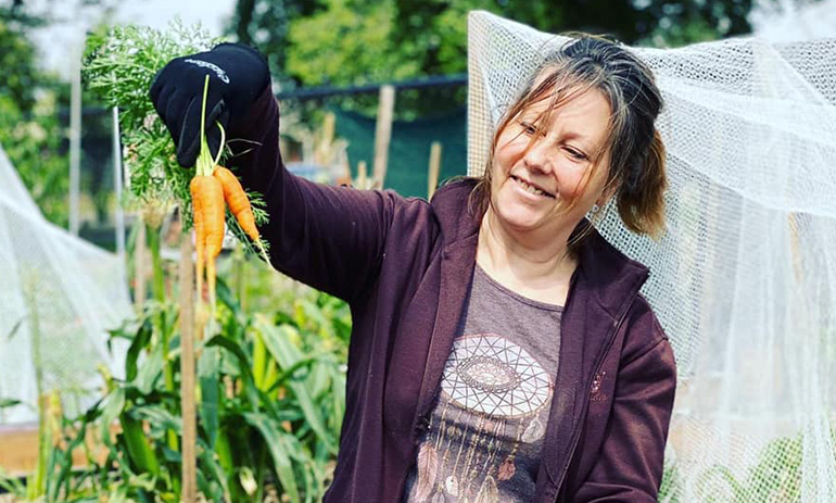 Lou Ridsdale, founder of Food is Free, proudly holds up a bunch of carrots. She is standing in a garden and is smiling at the carrots. She has her brown hair in a ponytail and is wearing a tshirt with a purple zip up hoodie over the top.