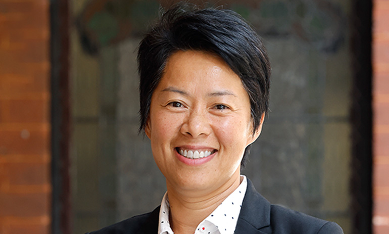 Hang Vo, a queer Asian woman, smiles at the camera. She is wearing a white collared shirt, dark blazer and has cropped black hair. We can only see her face and shoulders.