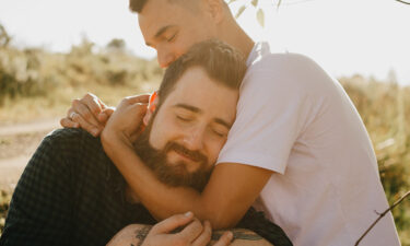 Two men hug in a field. One is wearing a black shirt and has a beard and dark hair; the other man is embracing his head and neck from the side and is wearing a white shirt.