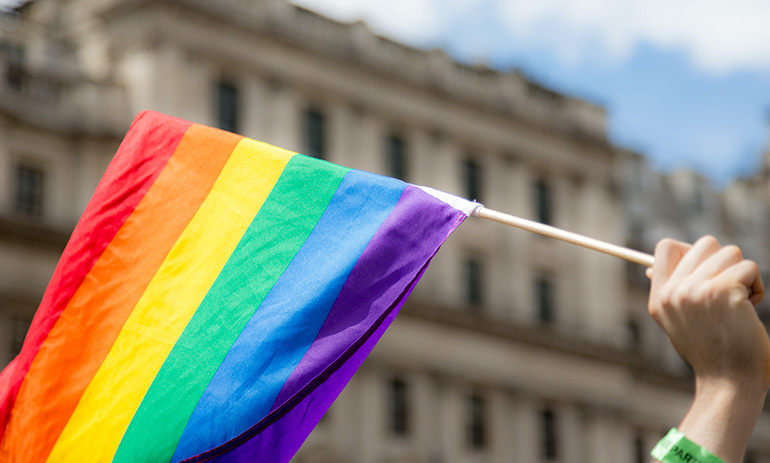 A rainbow Pride flag representing the LGBTQIA+ community, being waved in front of an old looking building.