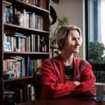 Ruby Susan Mountford sits at a table with their arms folded. They are wearing a red jumper and smiling up at the ceiling. There is a timber bookshelf behind them filled with books.