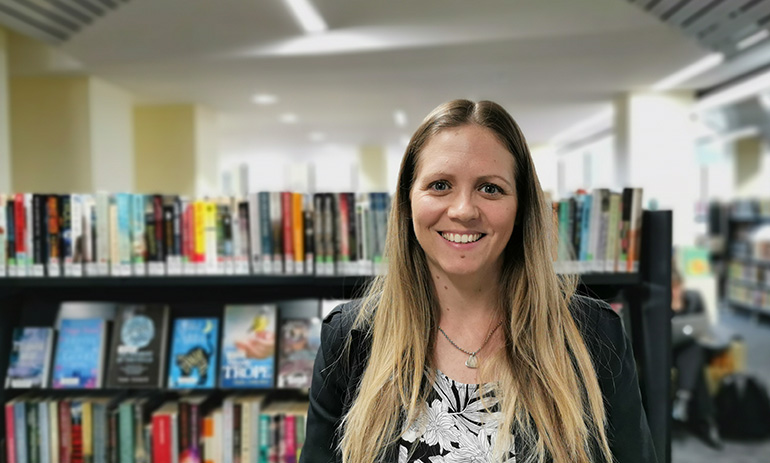Bianca Crocker smiles at the camera. She has long blonde hair and is white, and is wearing a white patterned top and a black blazer. She is standing in a library, surrounded by shelves of books.