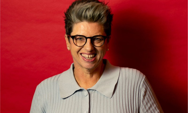 Nareen Young wears a grey ribbed shirt and black glasses, and smiles against a red background.